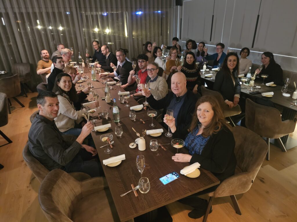 Two parallel tables in a restaurant space. People around the tables are looking at the camera, raising their glasses in a toast.