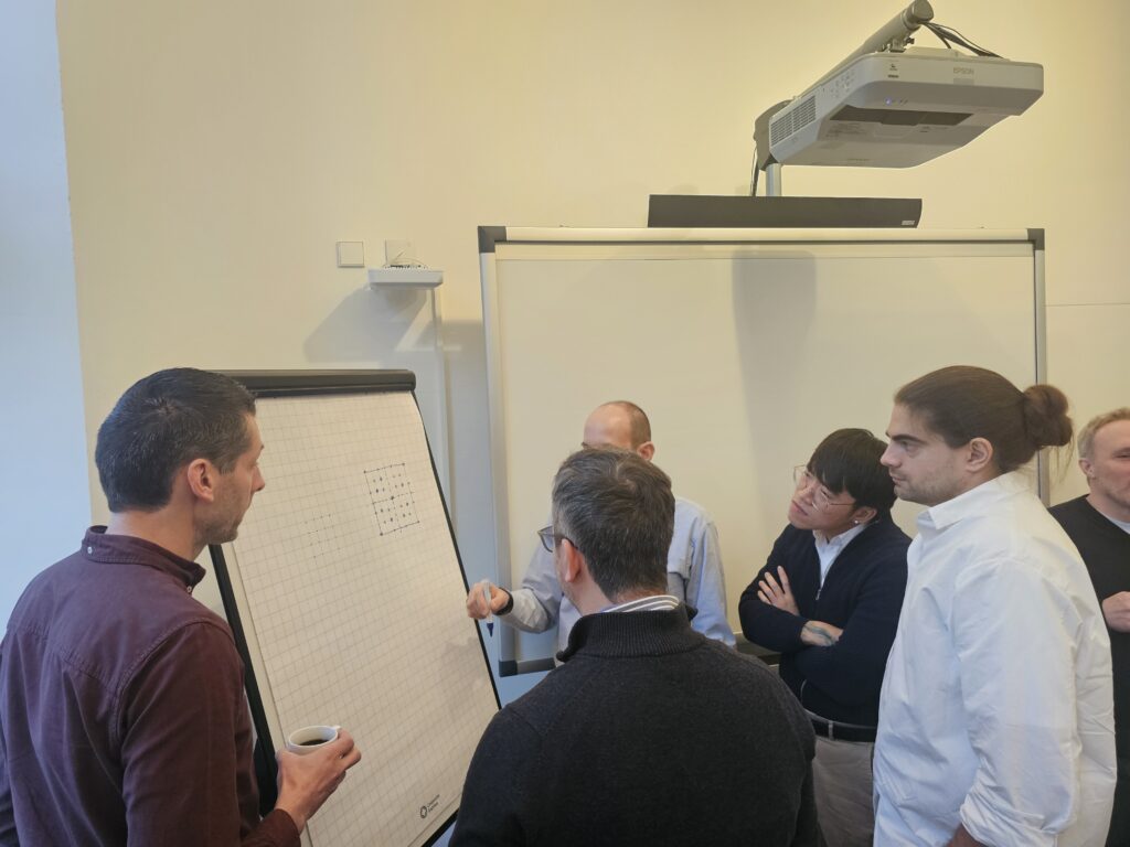 Four men stand in front of a flipchart in a white room, discussing sketches drawn on the paper by one of the men.