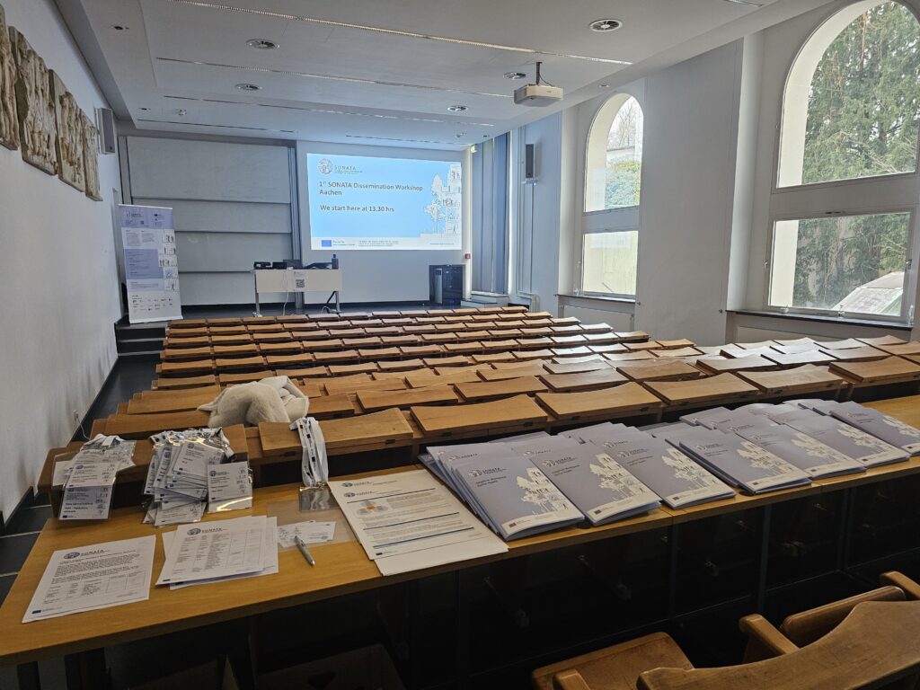 Classroom with rows of old-fashioned wooden seats and bench desks. A small stage at the front of the room has a SONATA banner, a desk and behind a presentation screen. In the foreground is SONATA materials, including a pile of lanyards and name tags, papers and seven rows of stacked blue folders with SONATA branding.