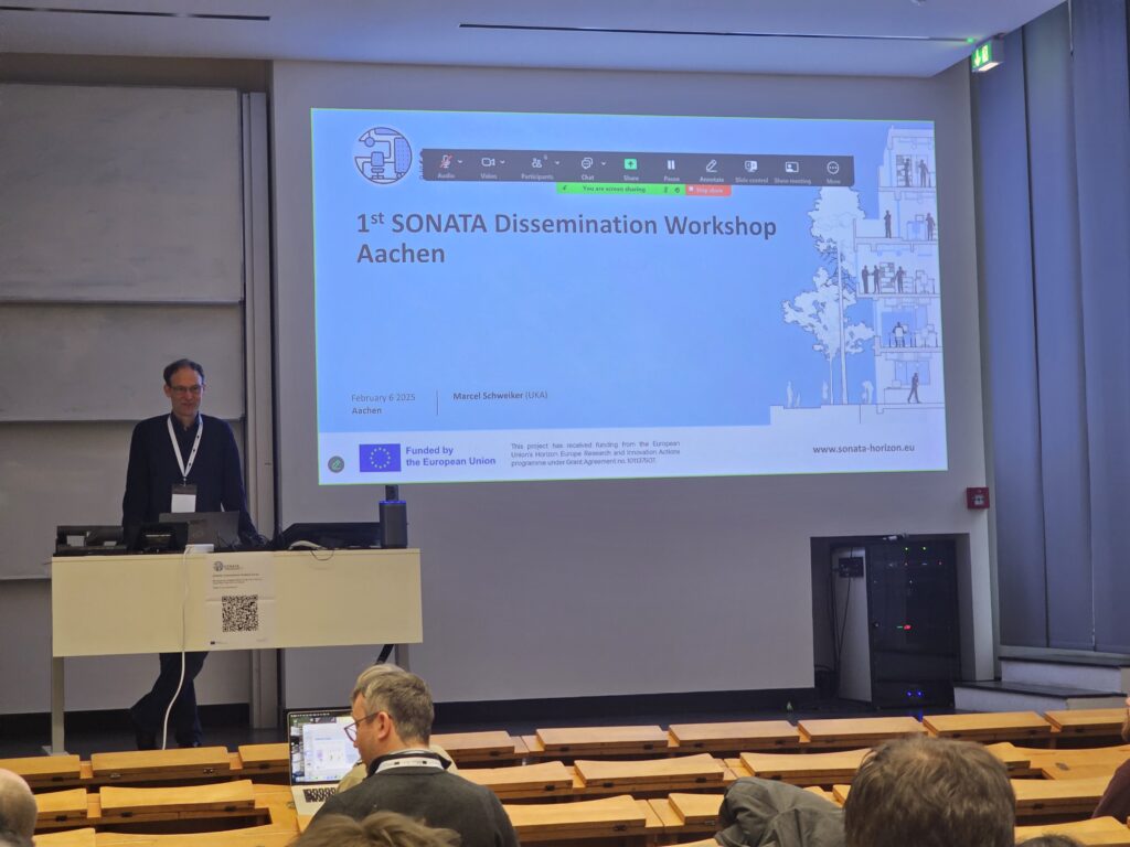 Man stands on a stage at the front of a classroom with rows of wooden seating and bench desks in the foreground. He stands behind a desk in front of a presentation screen with a slide reading "1st SONATA Dissemination Workshop Aachen".