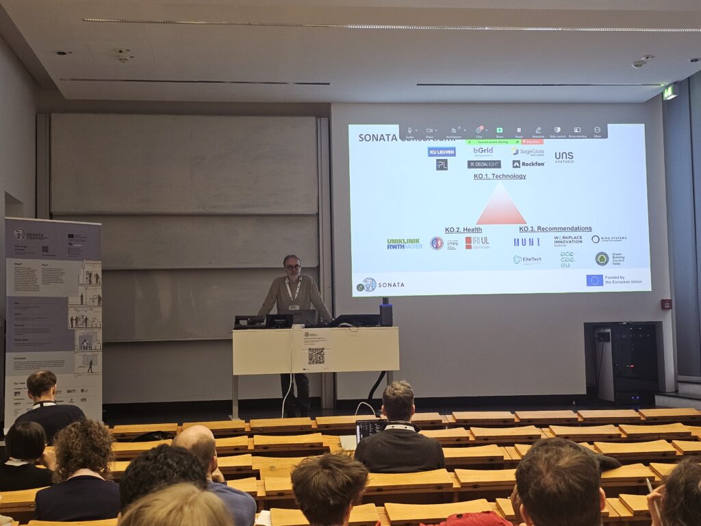 Man stands on a stage at the front of a classroom with rows of wooden seating and bench desks in the foreground. He stands behind a desk in front of a presentation screen with a slide containing project partner logos.