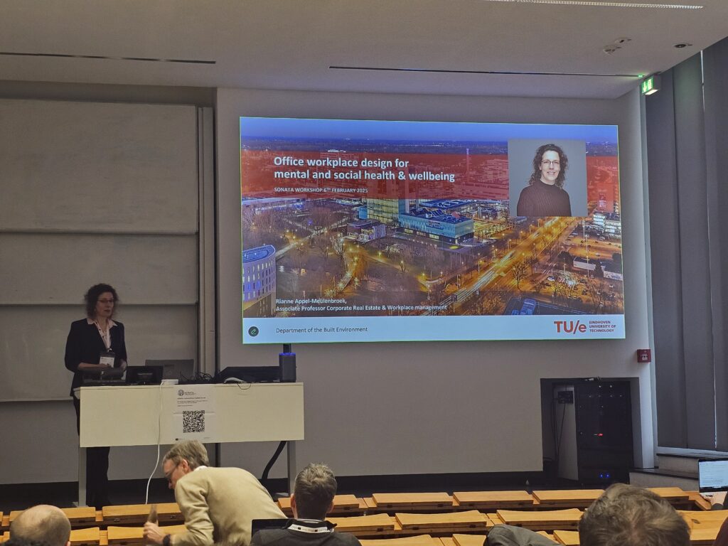 Woman stands on a stage at the front of a classroom with rows of wooden seating and bench desks in the foreground. She stands behind a desk in front of a presentation screen with a slide showing title "Office workplace design for mental and social health & wellbeing".
