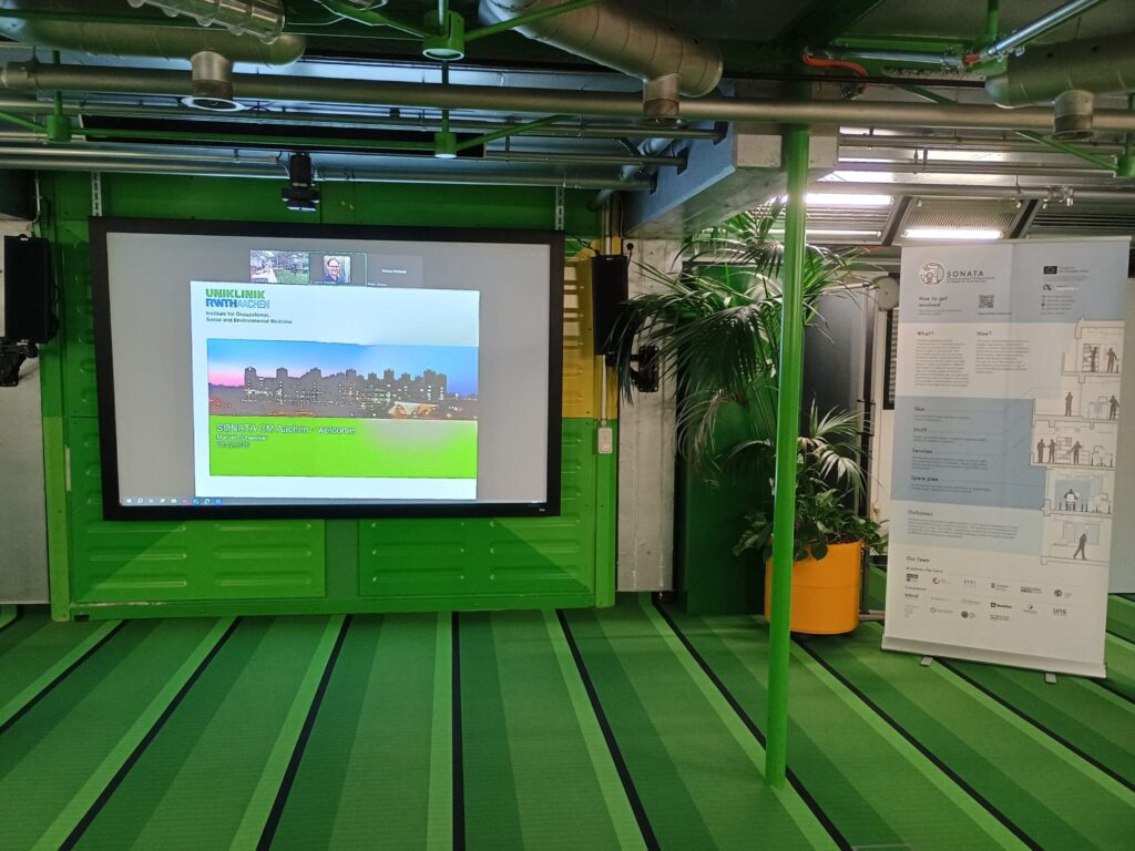 Meeting Room with presentation screen displaying Meeting Intro slide, SONATA banner poster stands to the right. Room has green decor, including green striped carpet, potted plants and exposed pipes suspended from the ceiling.