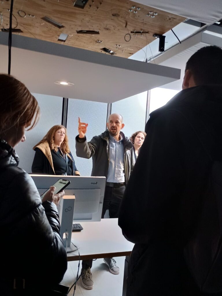 Small grey room with a panel, including a central light, suspended from the ceiling above a desk with computer and five people standing around the desk, one man points towards the lighting.