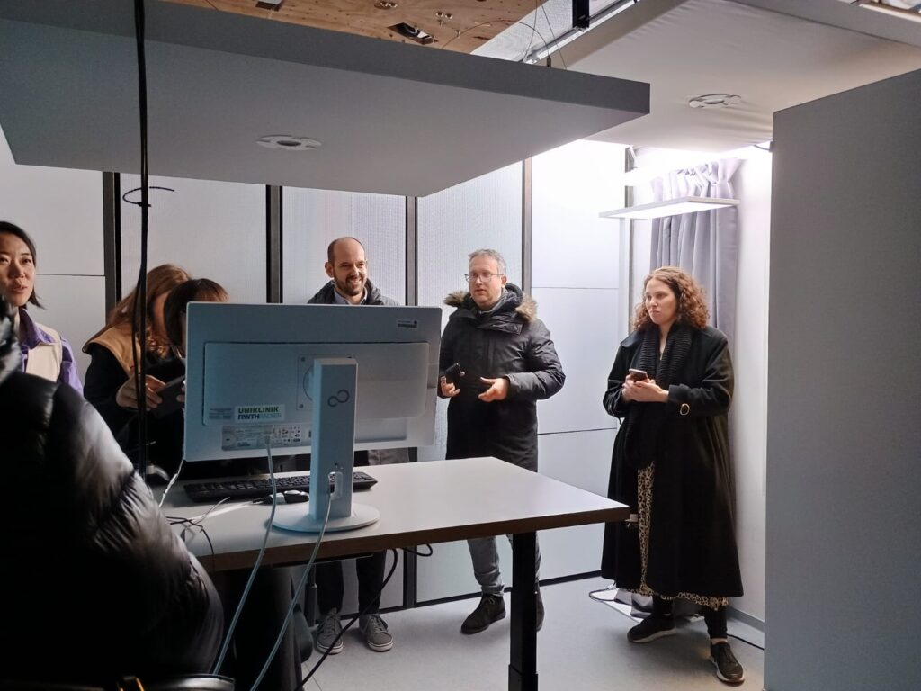 Small grey room with a panel, including a central light, suspended from the ceiling above a desk with computer and four people standing around the desk talking.