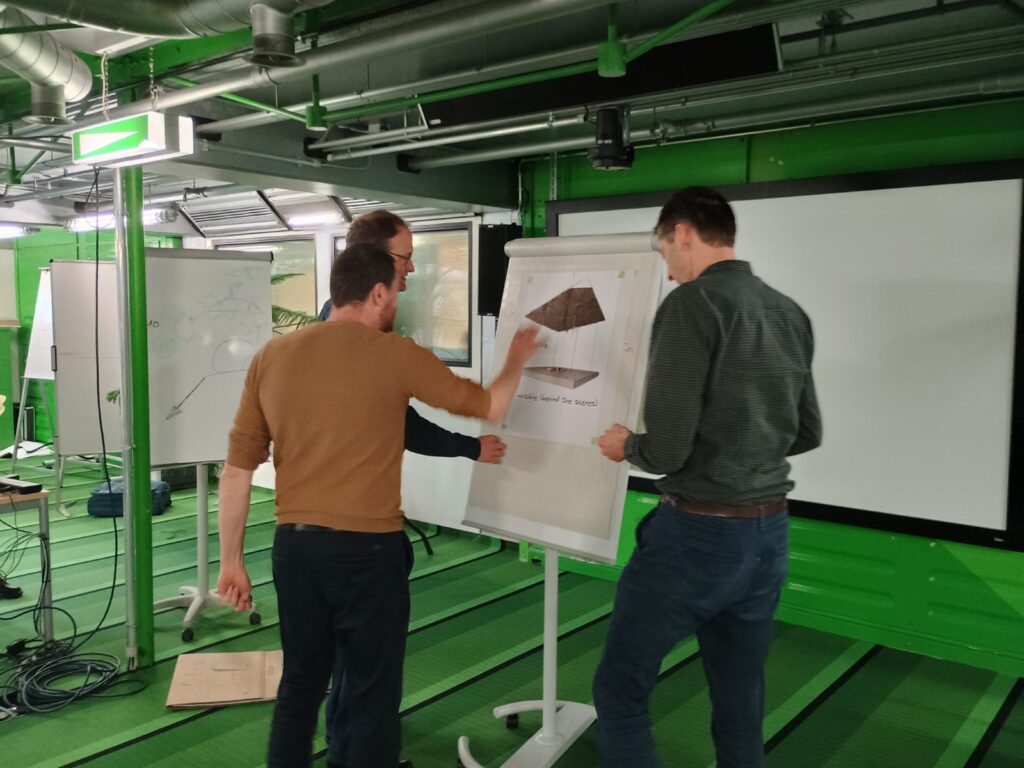 Three men stand in front of a flipchart, in a green meeting room, discussing an illustration pinned to he board, attaching post-it notes to the board.