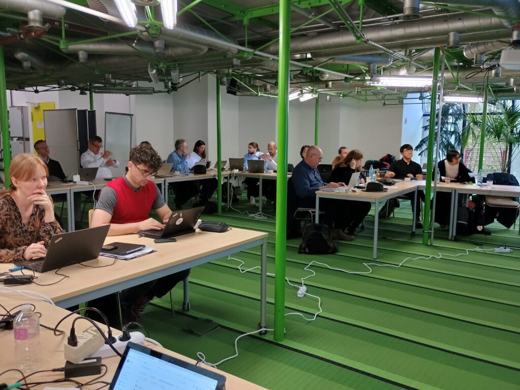 U-shaped arrangement of desks, with additional row in the middle with people sitting at desks with laptops in a green meeting space with exposed metal pipes suspended from the ceiling and green striped carpets.