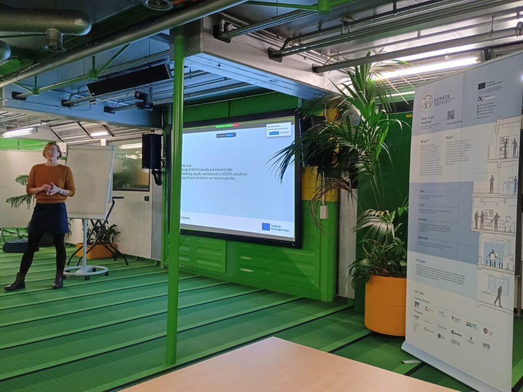 Woman stands in front of presentation screen, presenting (text on slide illegible) in a green meeting room. SONATA banner poster stands to the right of the picture.