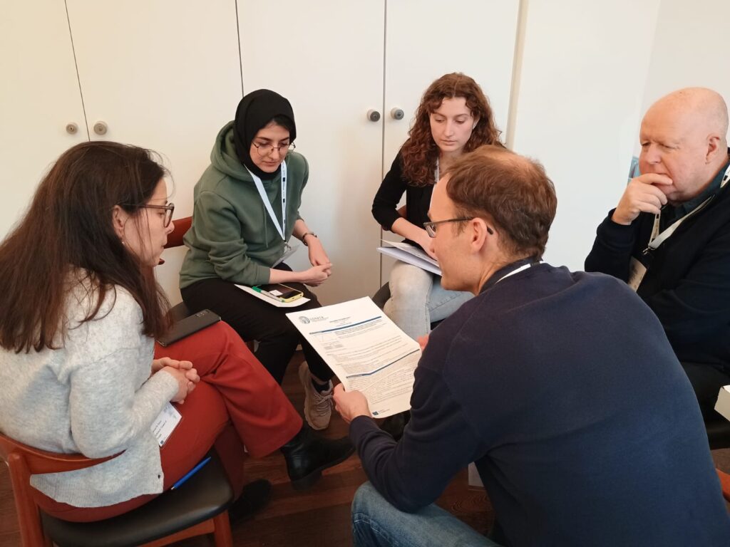 Five people sitting on chairs in a circle in conversation. One person holds a piece of white paper.