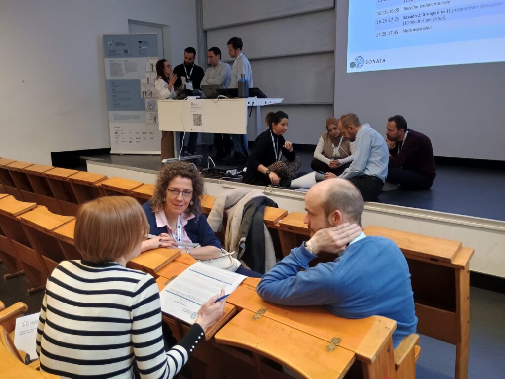 Classroom with group of three people sitting across two rows of wooden seating, in discussion in the foreground, on a stage in the background is one group of four people standing, talking around a desk and another group of four people sitting on the stage in a circle, talking.