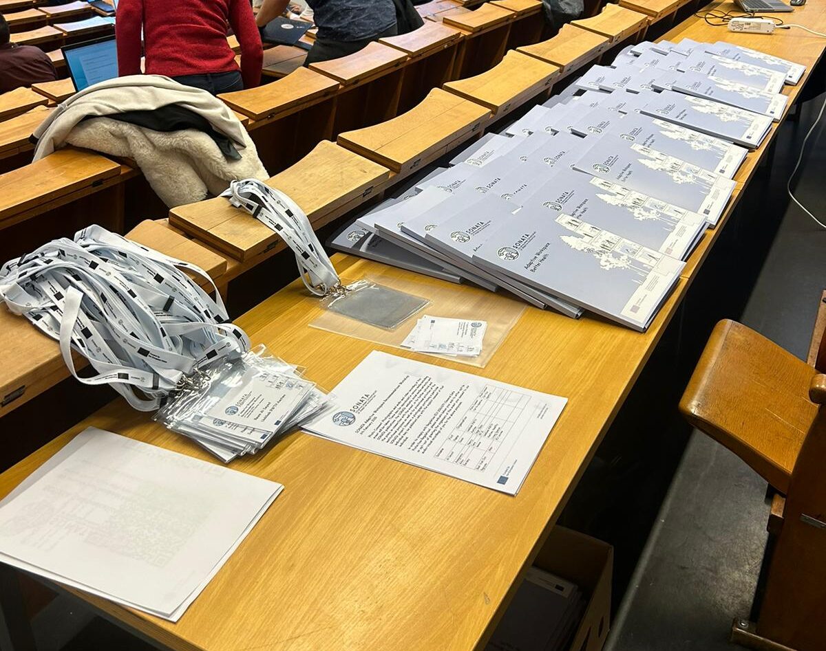 SONATA Recommendations Workshop A long wooden desk with collapsible wooden seating in front, in a classroom. On the desk are lanyards and name tags, registration sheets and blue SONATA-branded folders stacked and lined up in six parallel rows.