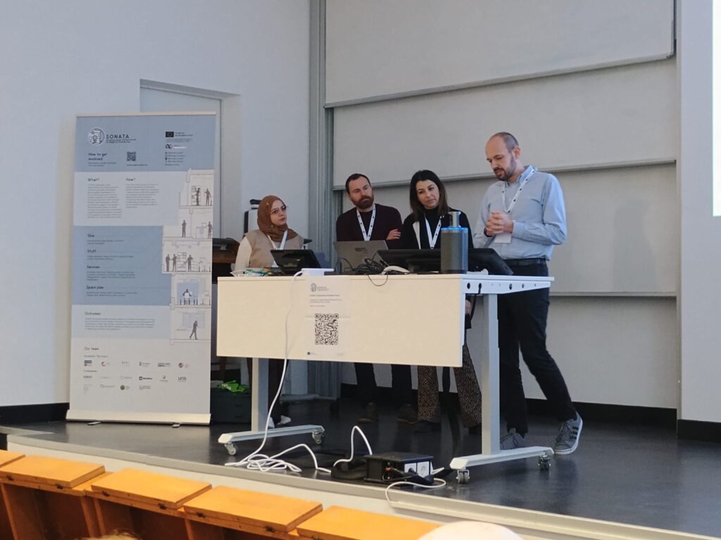 Four people stand behind a desk on a stage at the front of a classroom, one man talking. The SONATA banner poster sits to the left of the desk.