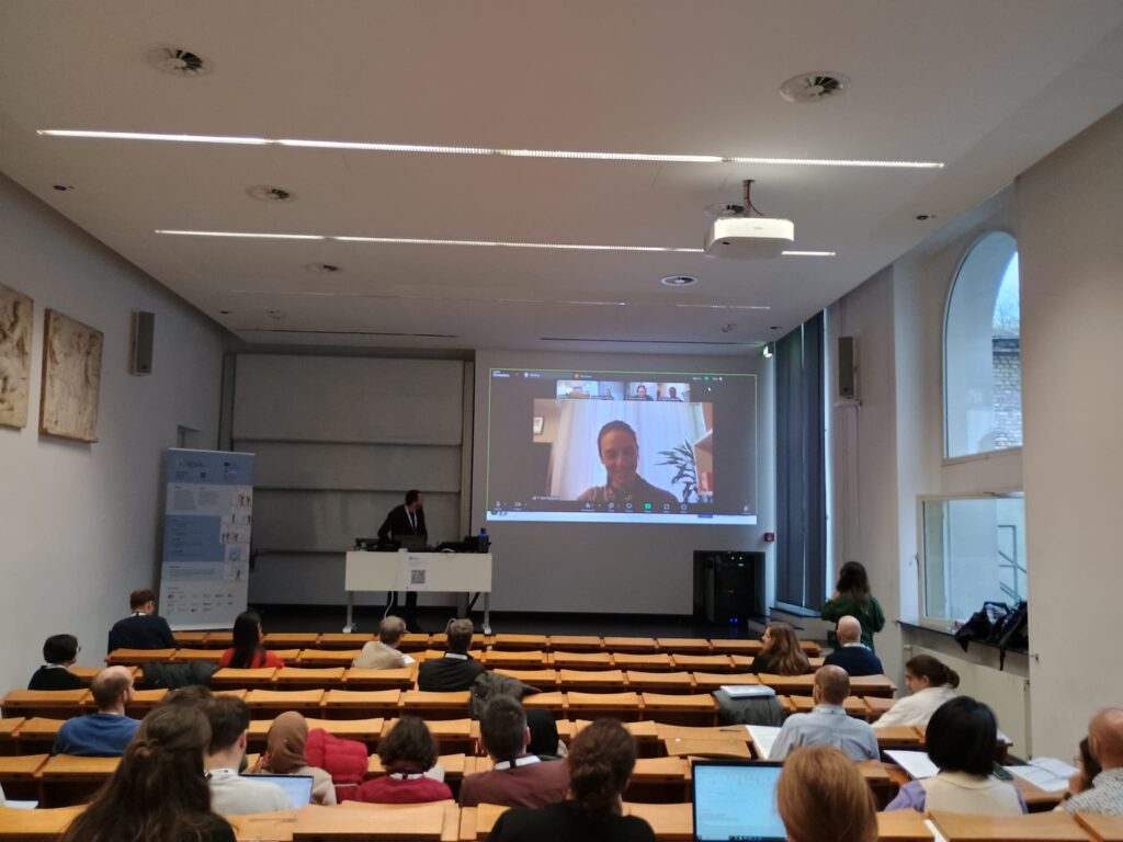 A classroom with rows of wooden seating and bench desks in the foreground, with people sitting scattered through the seating. A man stands behind a desk in front of a presentation screen displaying the video connection from an online attendee.