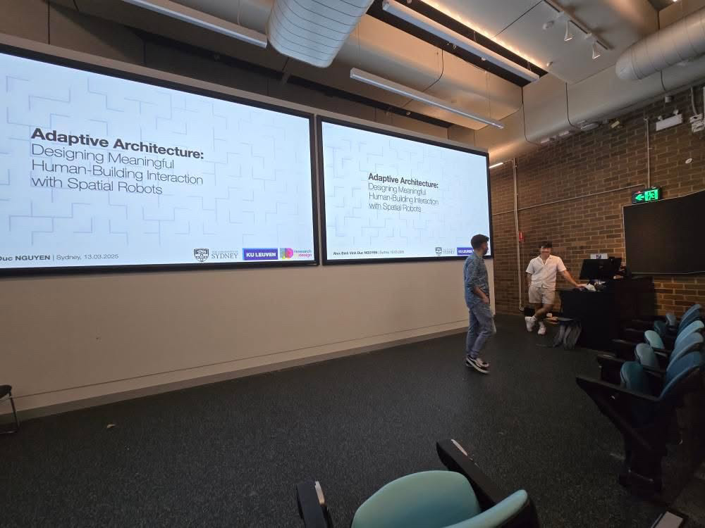 Large lecture room with two screens displaying "Adaptive Architecture: Designing Meaningful Human-Building Interaction with Spatial Robots." Two people stand near a desk.