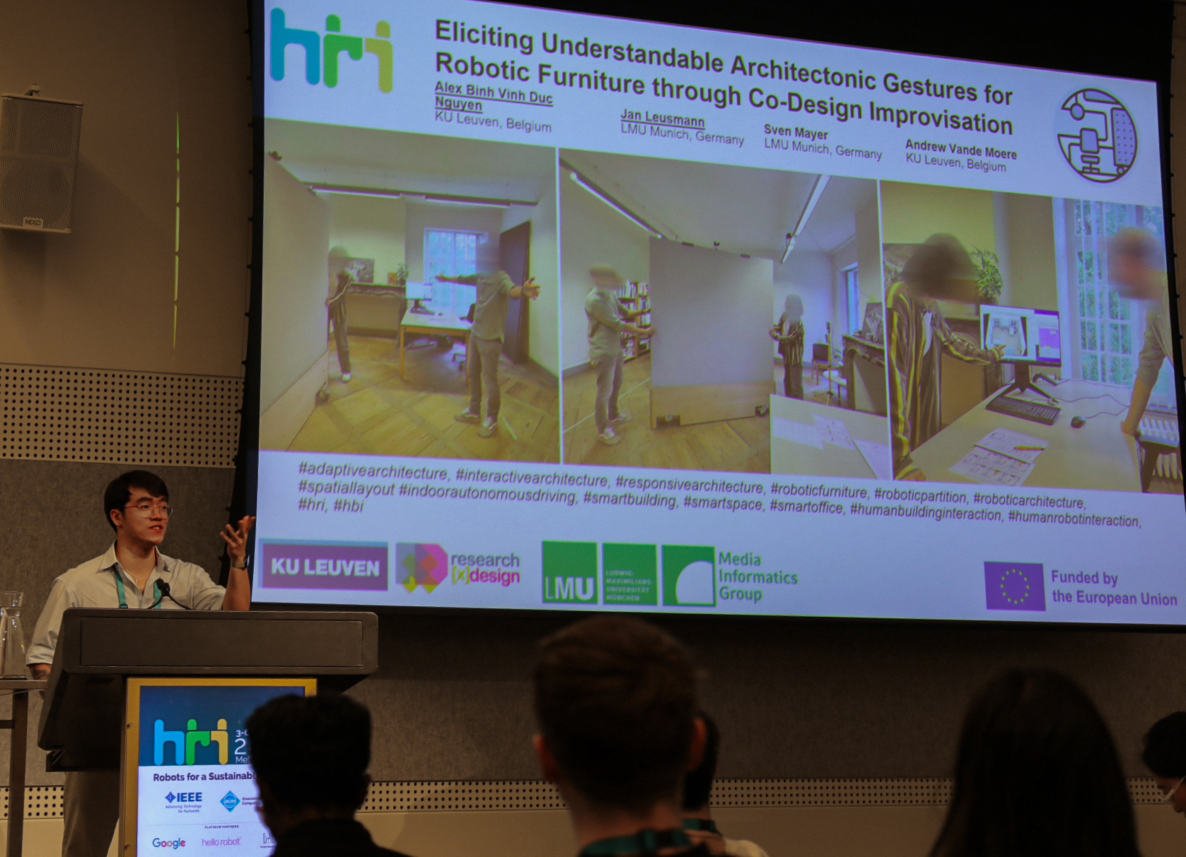A presenter at a podium speaks before a presentation slide titled "Eliciting Understandable Architectonic Gestures for Robotic Furniture through Co-Design Improvisation." The slide features images of people interacting with robotic partitions in a bright room. Logos of universities and research groups are displayed below, indicating collaboration and EU funding. The atmosphere is professional and academic.