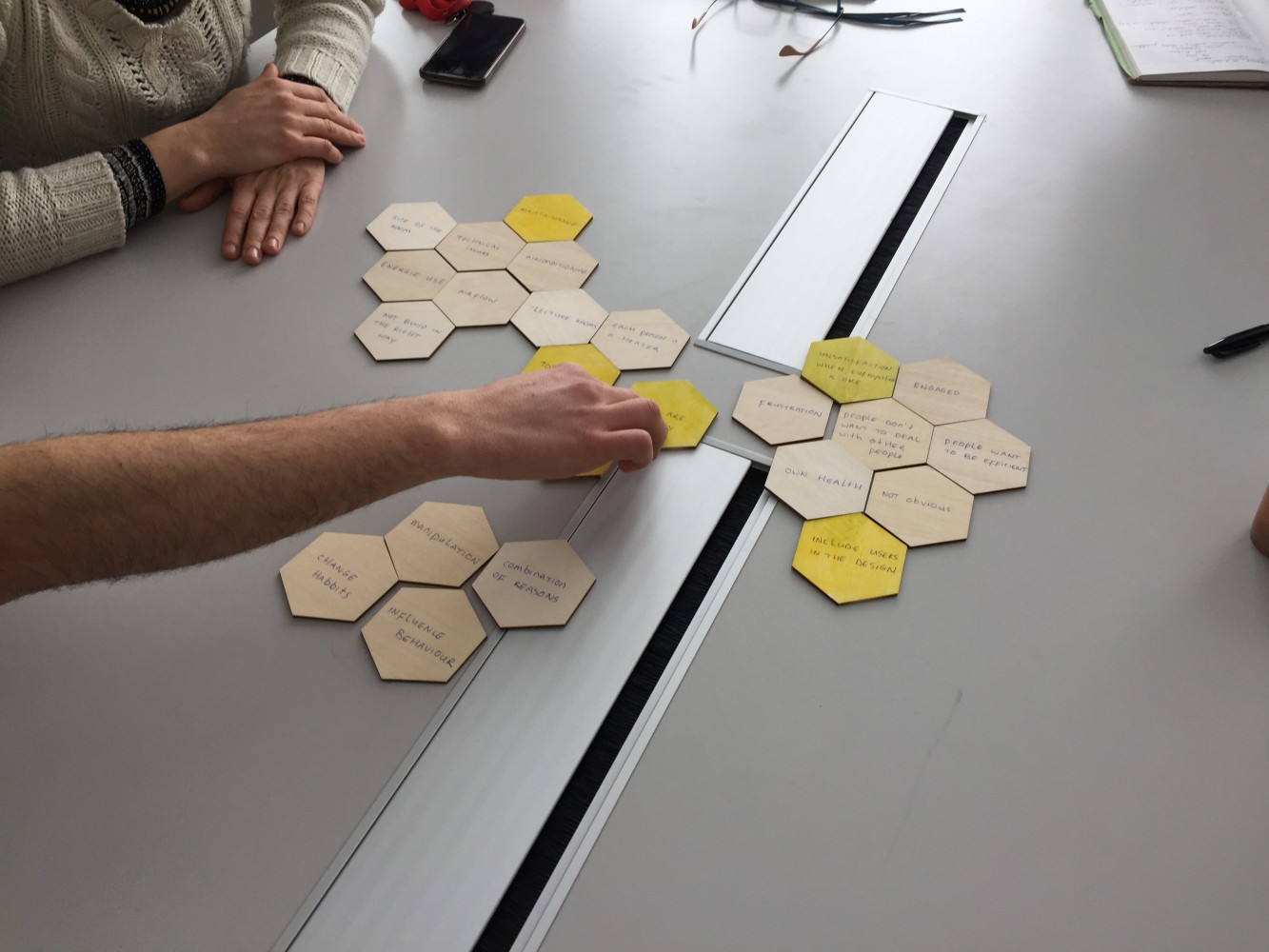 Two people engage in a collaborative activity, arranging hexagonal tiles with written notes on a table. The scene is studious and focused.