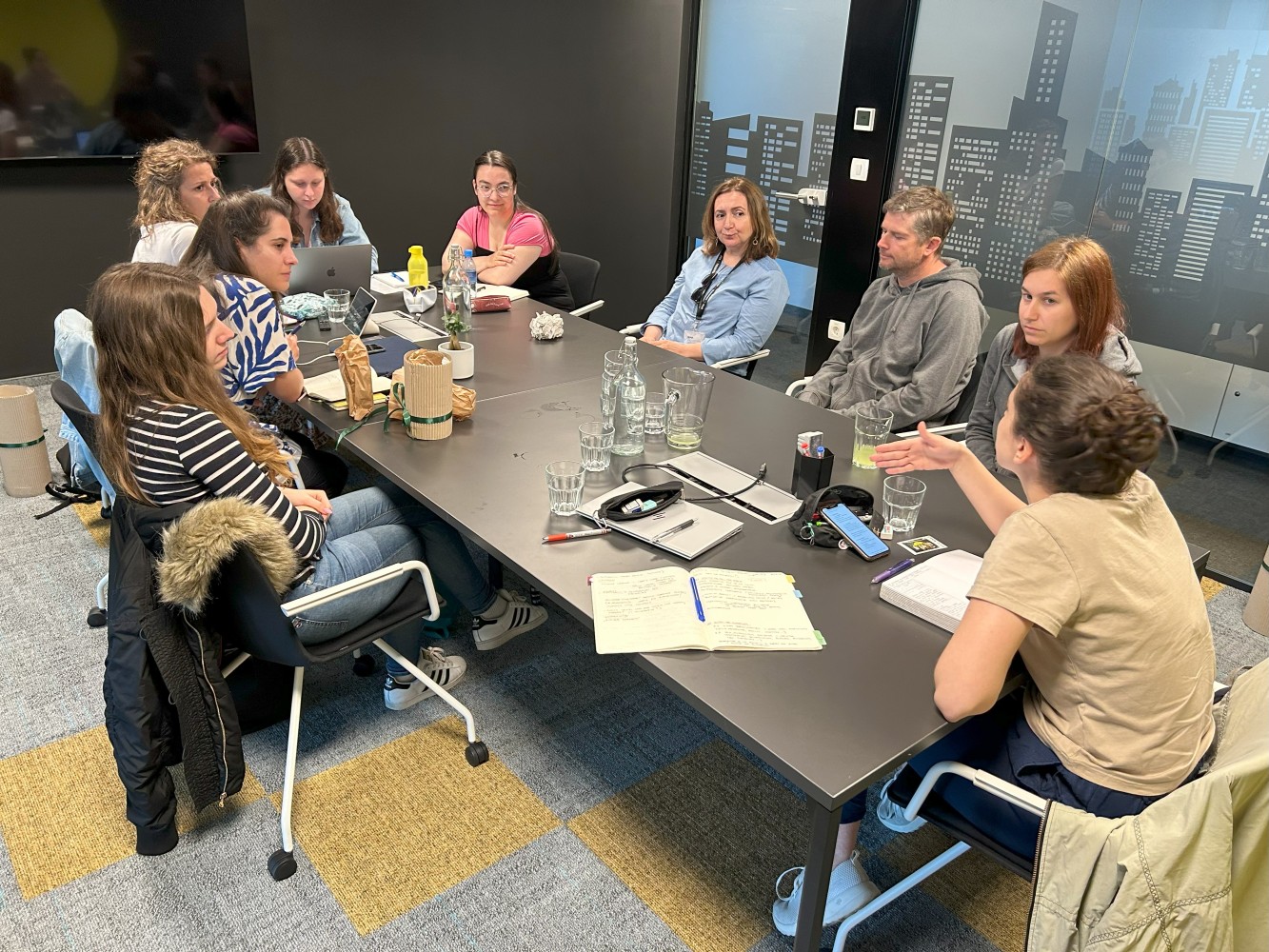 A group of nine people sit around a conference table in an office, engaged in discussion. Notebooks, a laptop, and glasses are scattered on the table. The room has a cityscape mural and a collaborative atmosphere.