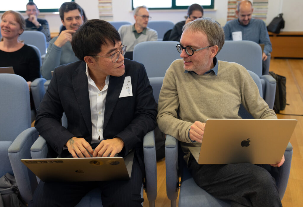 Two men sitting in blue chairs at a conference, holding open laptops and engaged in conversation, while others in the background are smiling.