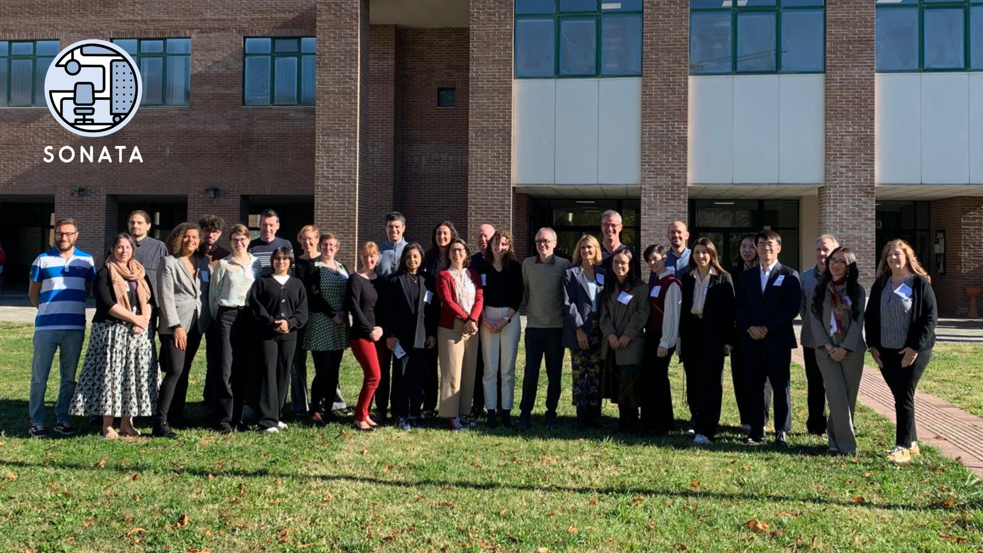 SONATA 6th Consortium Meeting A group of 29 people stands outside a red brick building on a sunny day, smiling. The "SONATA" logo is in the top left corner. The mood is professional and cheerful.