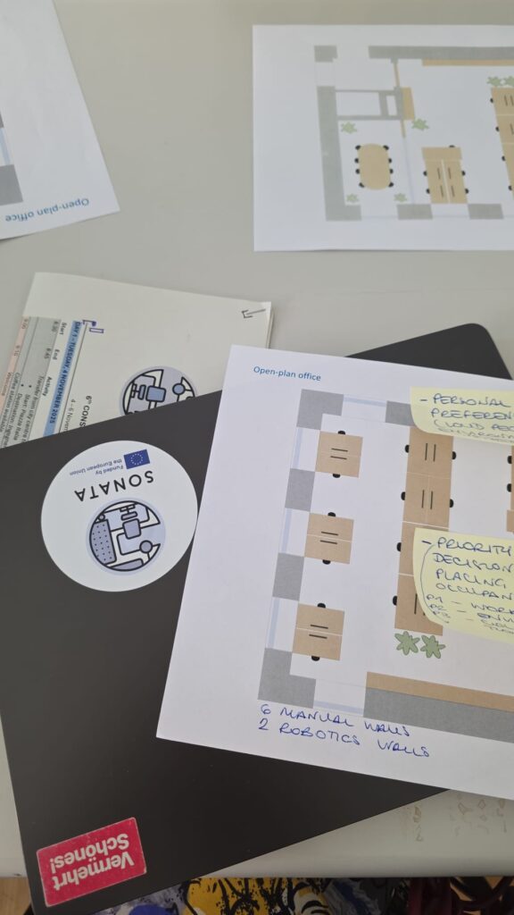 A desk holds a laptop with "SONATA" sticker, and papers showing an open-plan office design with handwritten notes and diagrams.