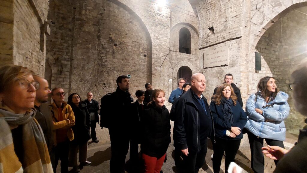 A group of people in winter clothing attentively listen inside a medieval stone chamber with arched ceilings, conveying curiosity and interest.