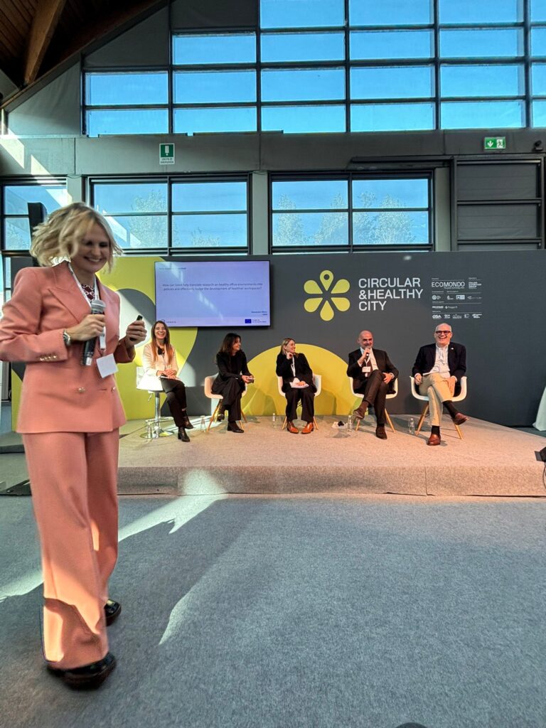 A woman in a pink suit walks with a microphone in a bright conference hall. Behind her, a panel of six people sits with a sign reading "Circular & Healthy City."