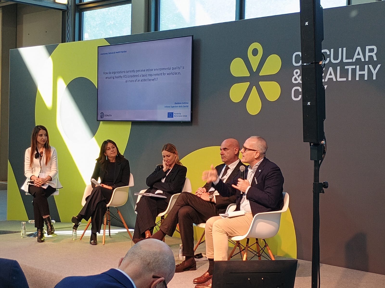 Panel of five people seated in a modern conference room with a bold green backdrop and screen. They engage in discussion, conveying a collaborative and professional atmosphere. Keywords: conference, panel, discussion, environment, collaboration.