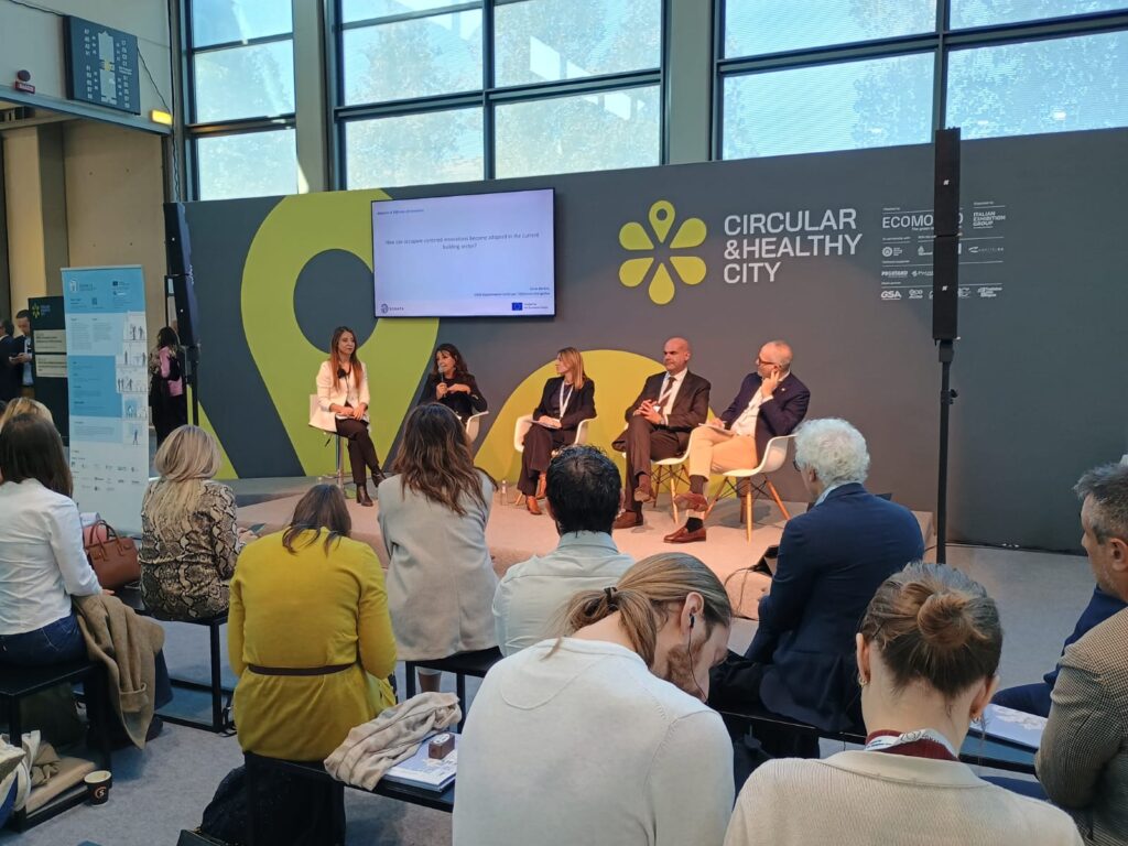 Panel discussion on "Circular & Healthy City" with four speakers seated, addressing an audience. Bright, spacious setting with a screen behind them.