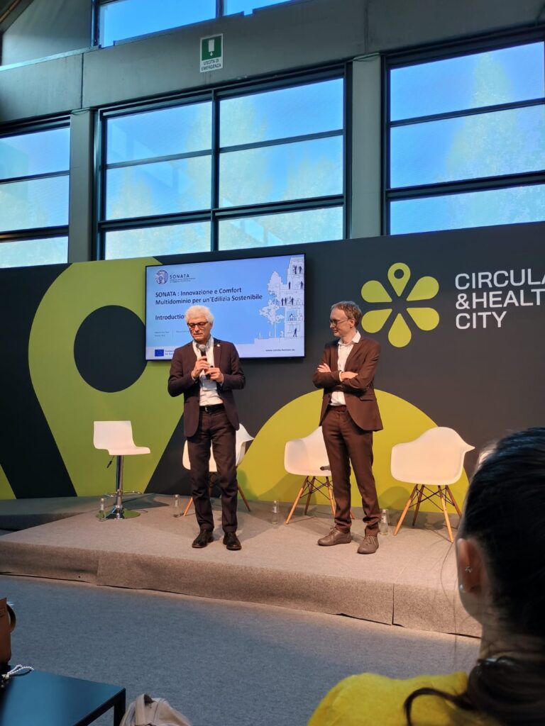 Two people in suits stand on stage with a green and black backdrop, featuring a logo and text, ``Circular & Healthy City.'' A presentation screen displays ``Innovazione e Comfort Multisensoriale'' in Italian.