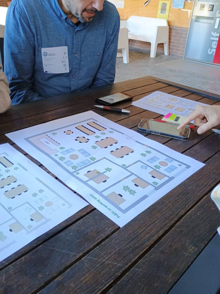 A man in a blue shirt sits at a wooden table, examining architectural floor plans with labeled rooms and plants. A hand points at a phone. The setting is informal and collaborative.