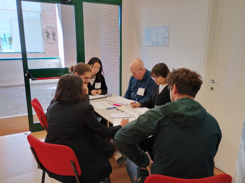 A group of six people sit around a small table in a meeting room. They focus on papers, writing and discussing, conveying a collaborative atmosphere.
